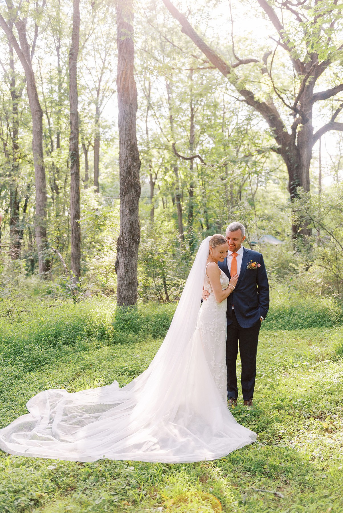 Bride and groom standing in a sunlit forest, bride in a long white gown, groom in a dark suit, trees in background.