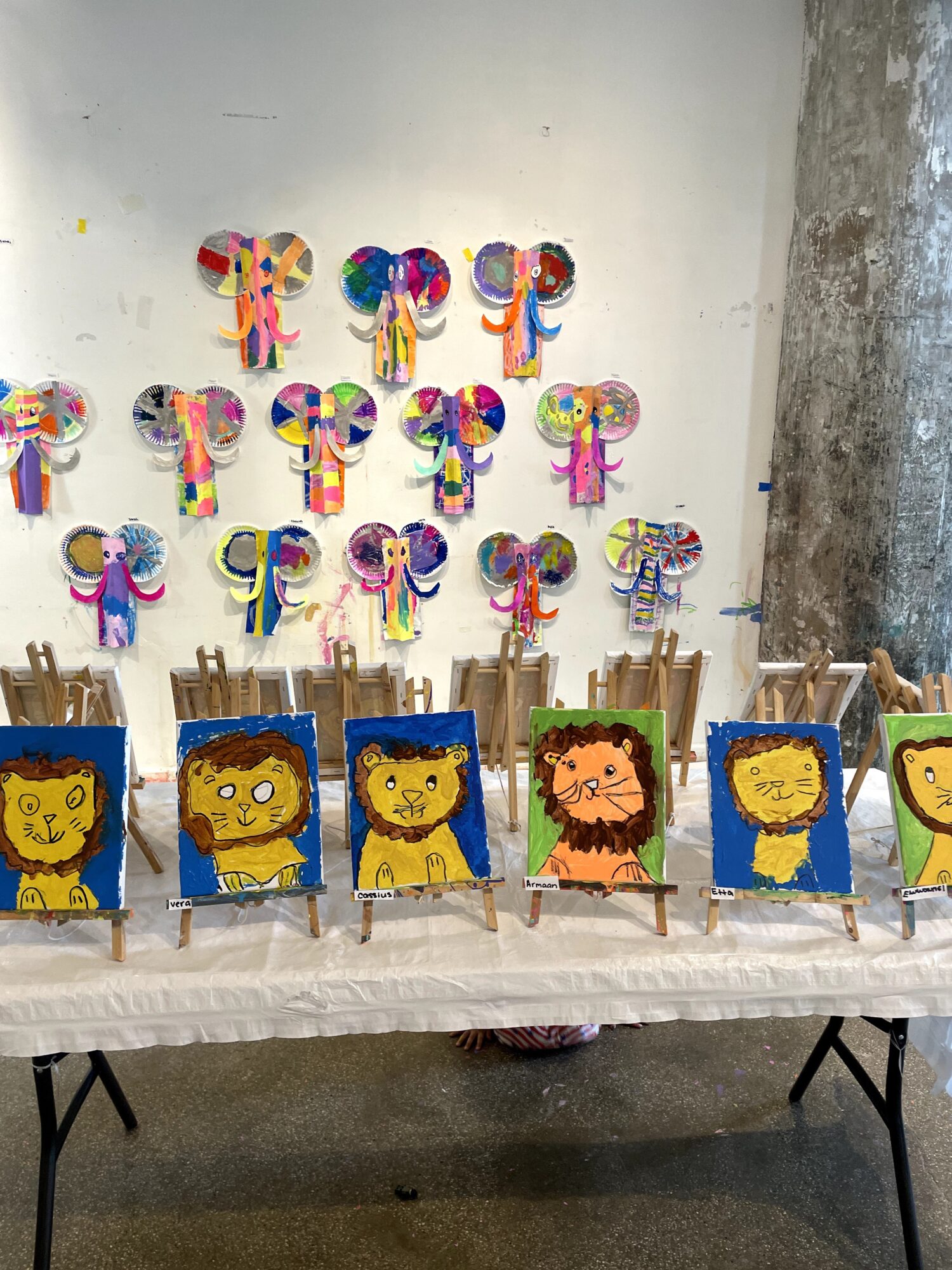 Table with lion-themed party hats and animal face decorations, wall with colorful paper animal crafts, plain wall and concrete pillar.