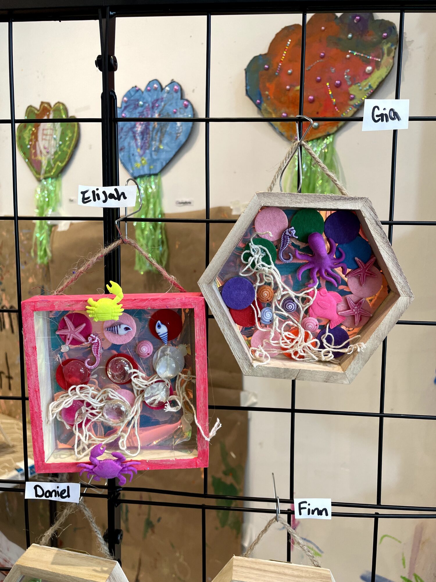 Colorful decorated cookies shaped like hearts and butterflies, with labeled tags for Elijah, Gina, Daniel, and Fim, displayed on a black grid.
