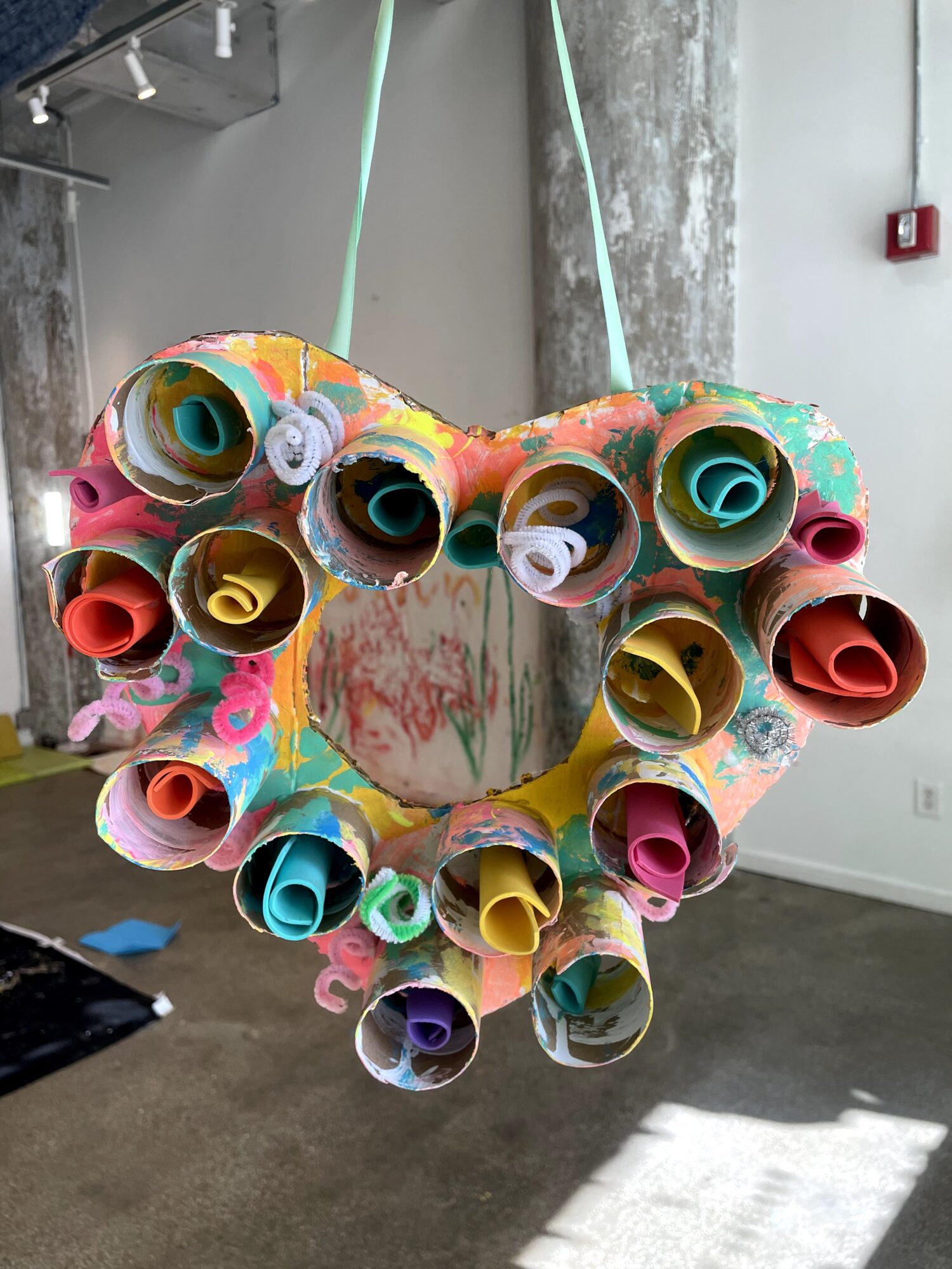 Colorful heart-shaped craft made of rolled paper tubes hanging indoors, with a concrete wall and window in the background.