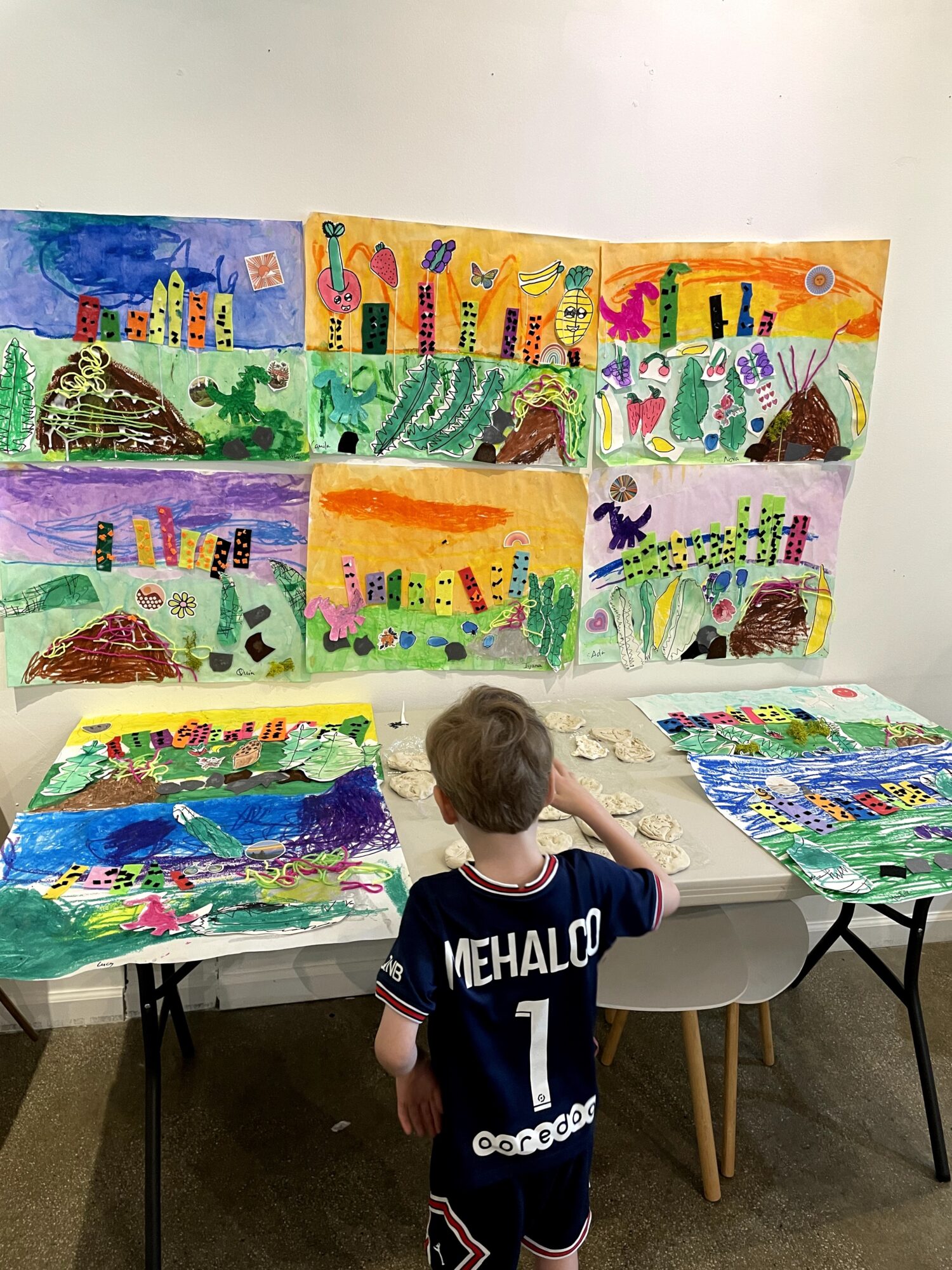 Child with back facing, wearing a black jersey, looking at colorful children's drawings on tables and wall.