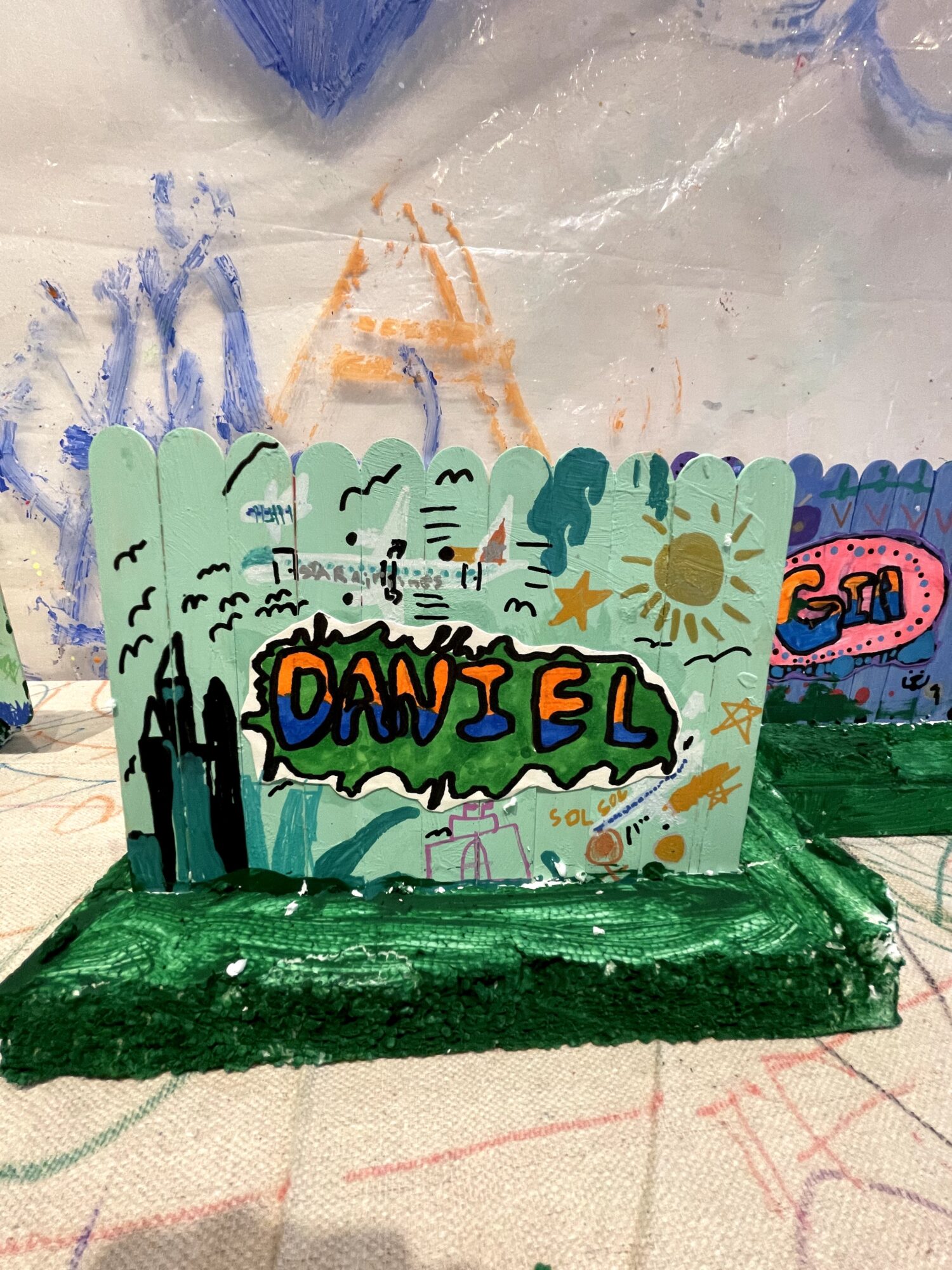 Colorful painted sign with the name 'DANIEL' in bold letters, surrounded by trees, sun, and abstract shapes, on a green base.