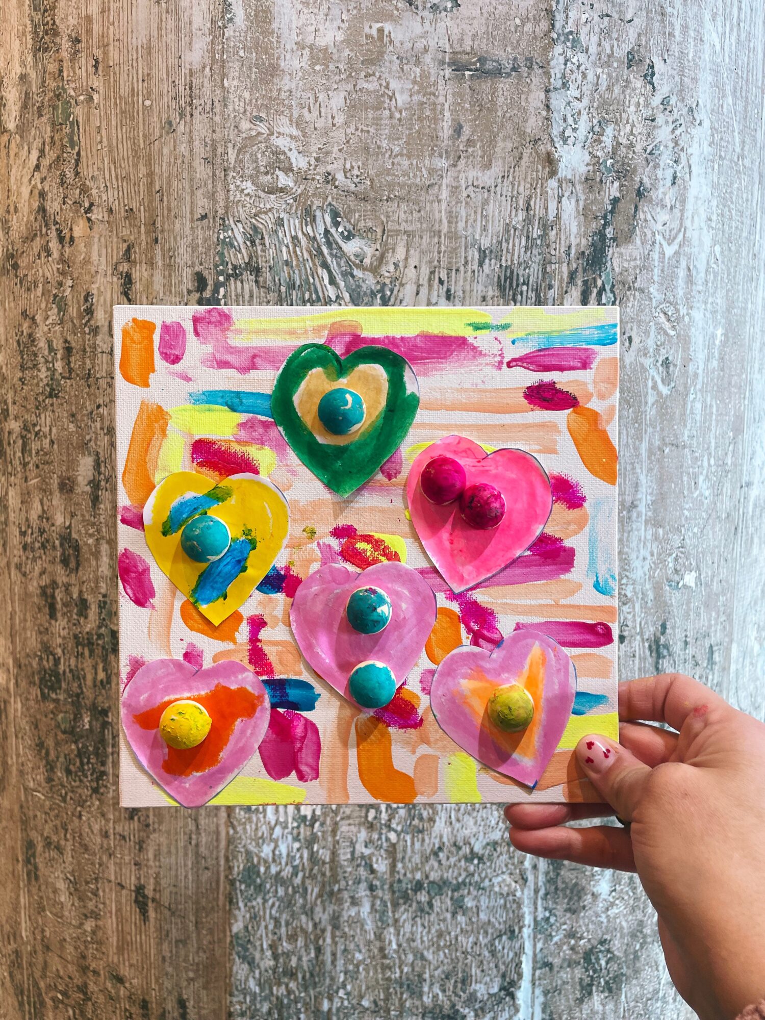 Child's hand holding colorful heart-shaped and round objects on a painted background.