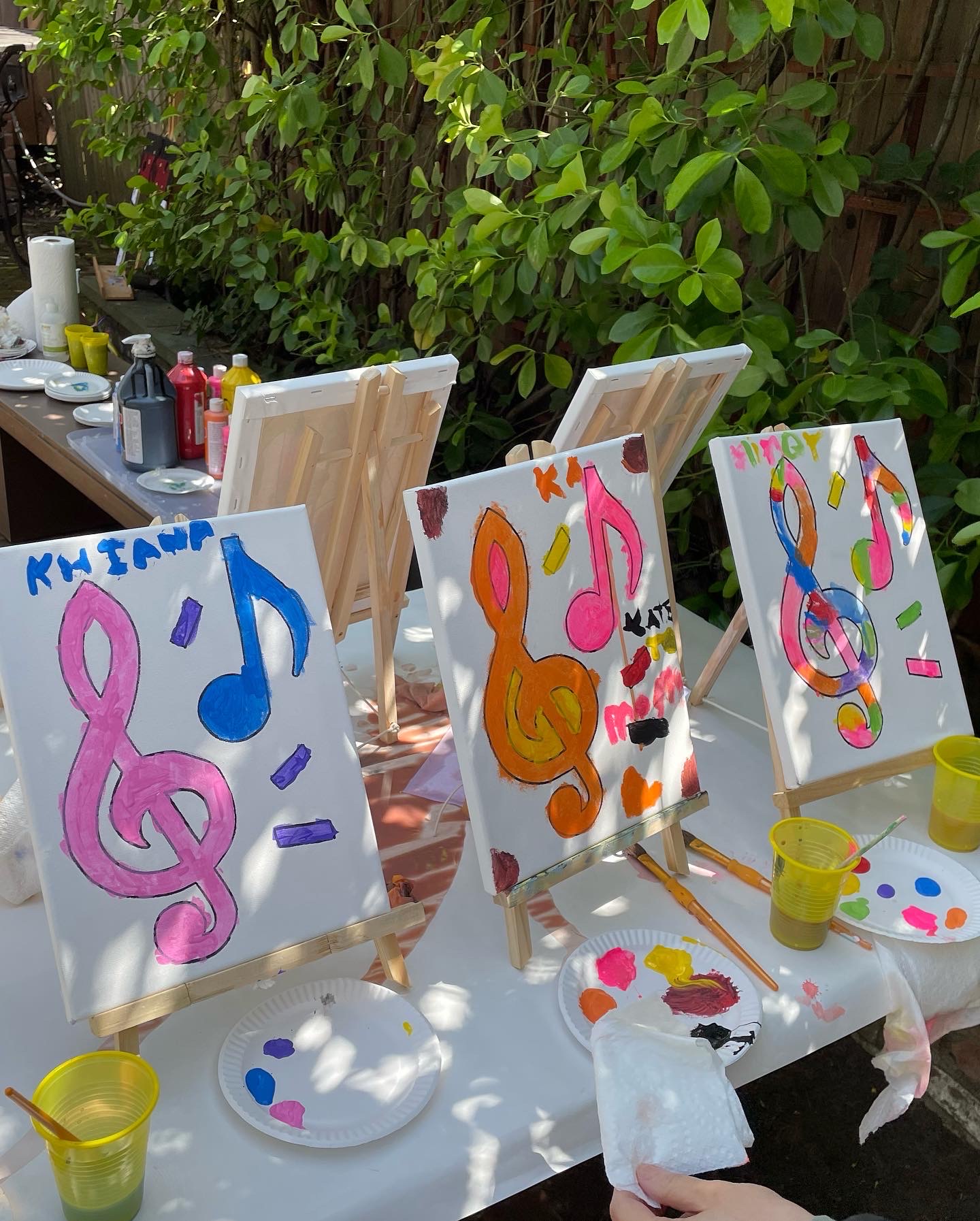 Three paintings on easels depict colorful musical symbols and abstract designs outdoors with art supplies nearby.