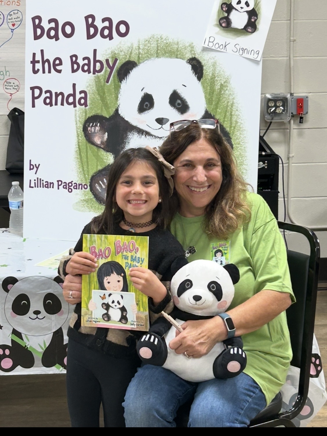 A woman and a girl sit together, holding a panda plush toy and a book, in front of a panda-themed display.