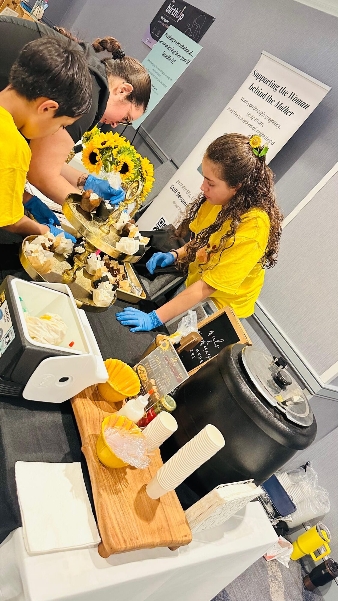 Children in yellow shirts gather around a table with a cake, flowers, and a rice cooker, in a room with informational signs.