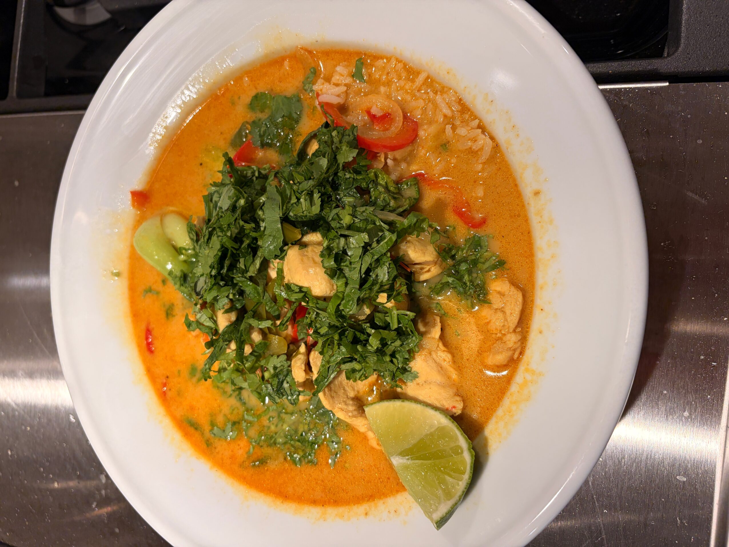 Bowl of orange-colored soup with herbs, vegetables, and a lime wedge, viewed from above.