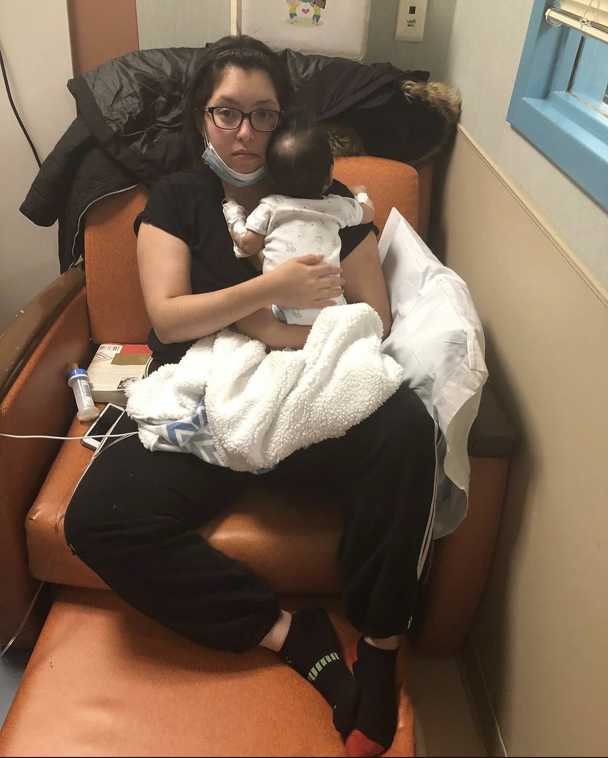 Woman sitting on hospital bed holding a baby, with medical supplies nearby, in a room with a window and blue trim.