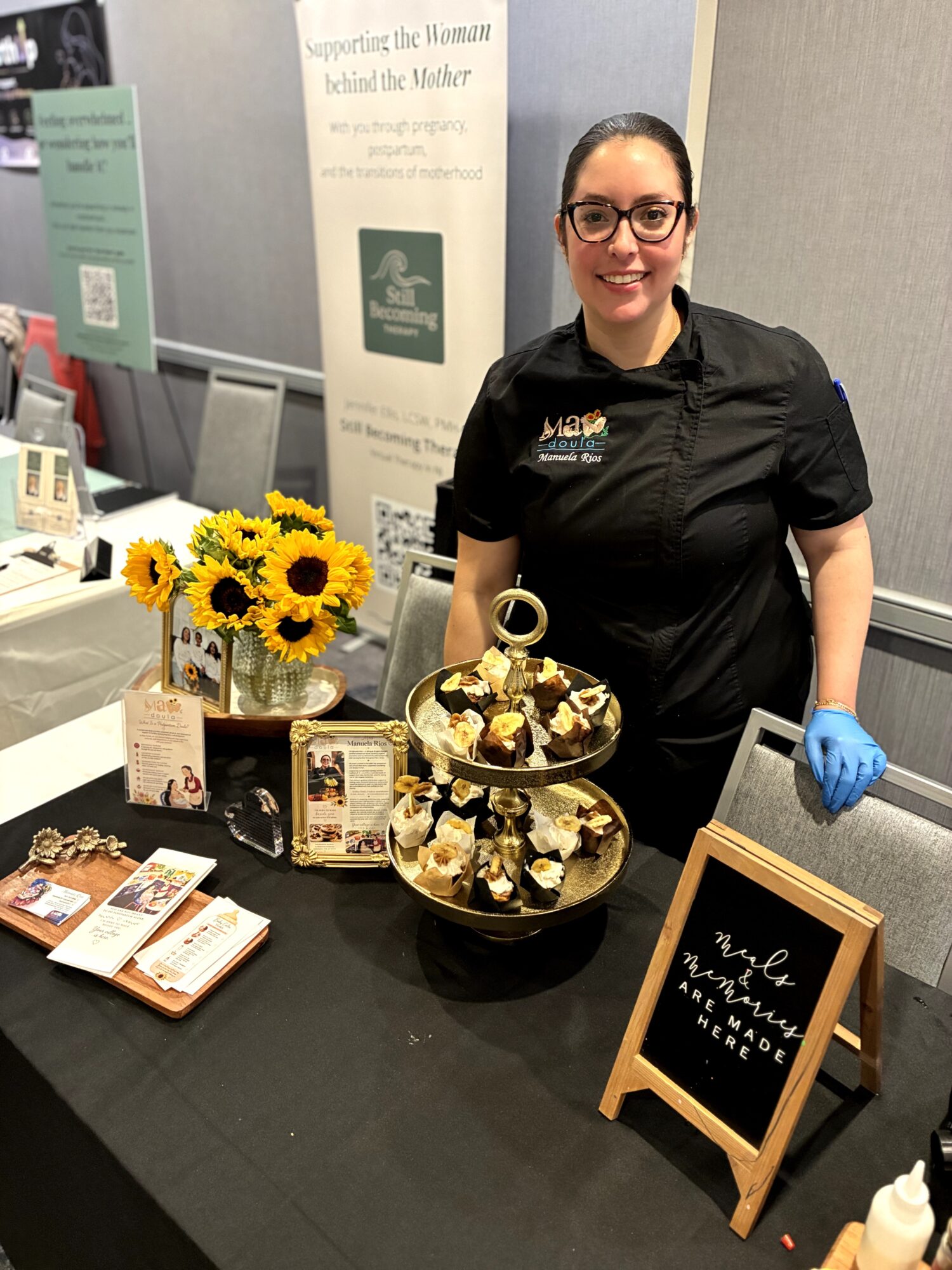 Smiling woman in black shirt and glasses stands behind a table with sunflower bouquet, chocolates, brochures, and a chalkboard sign.