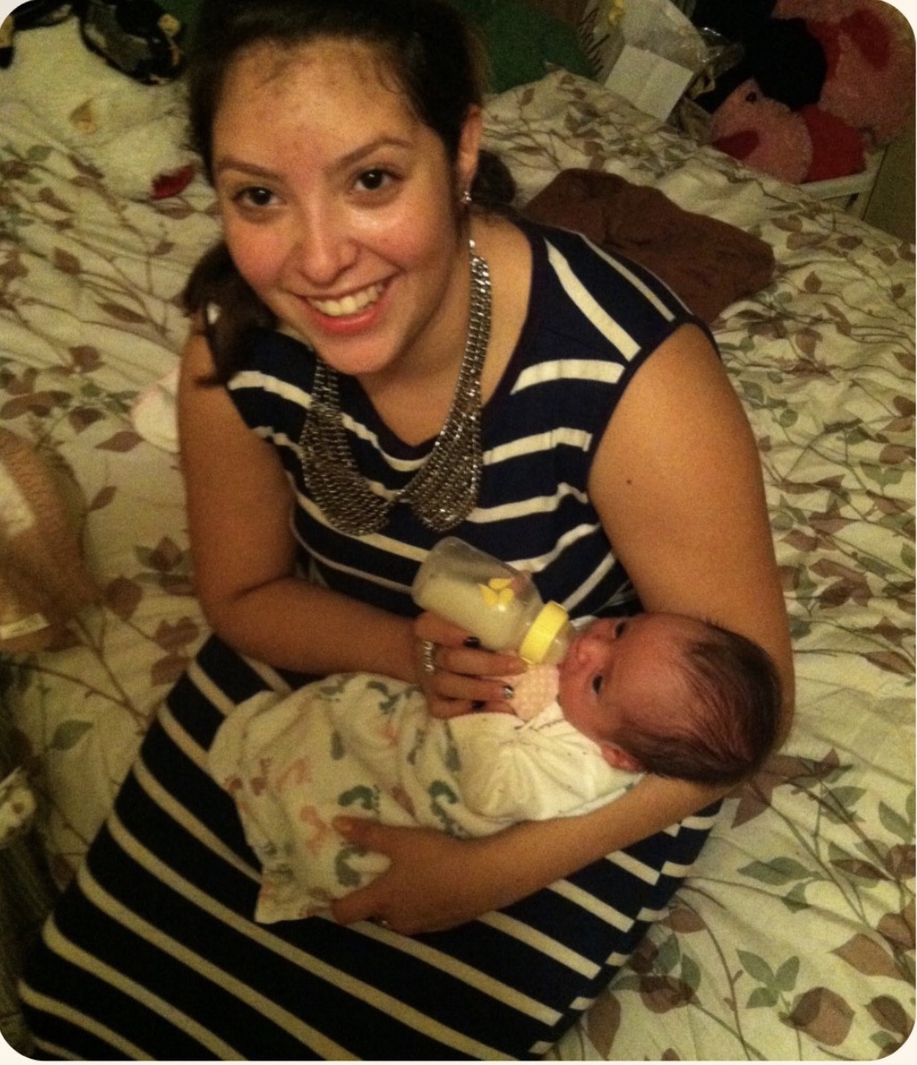 Young woman smiling and holding a baby bottle, cradling a newborn baby on a bed with floral sheets.
