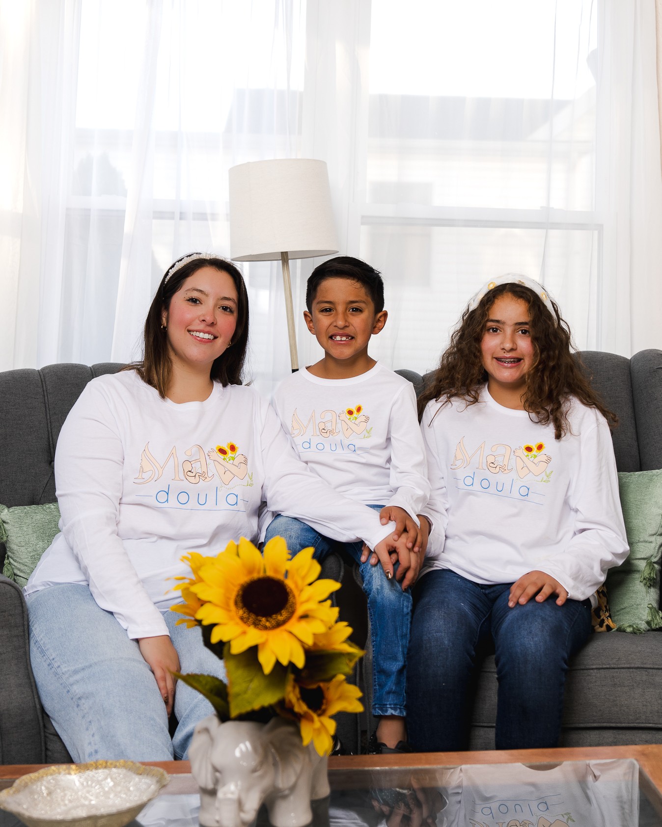 Three children sitting on a sofa, holding a sunflower, smiling, in a bright room with curtains and a lamp behind them.