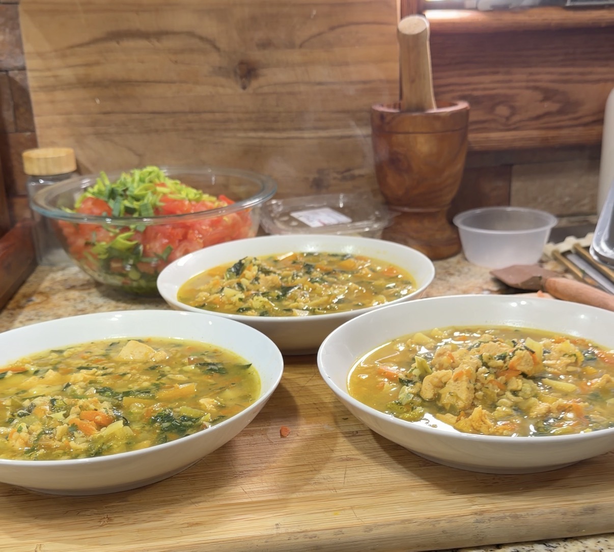 Three bowls of soup on a wooden table with a salad, a mortar and pestle, and a cup in the background.