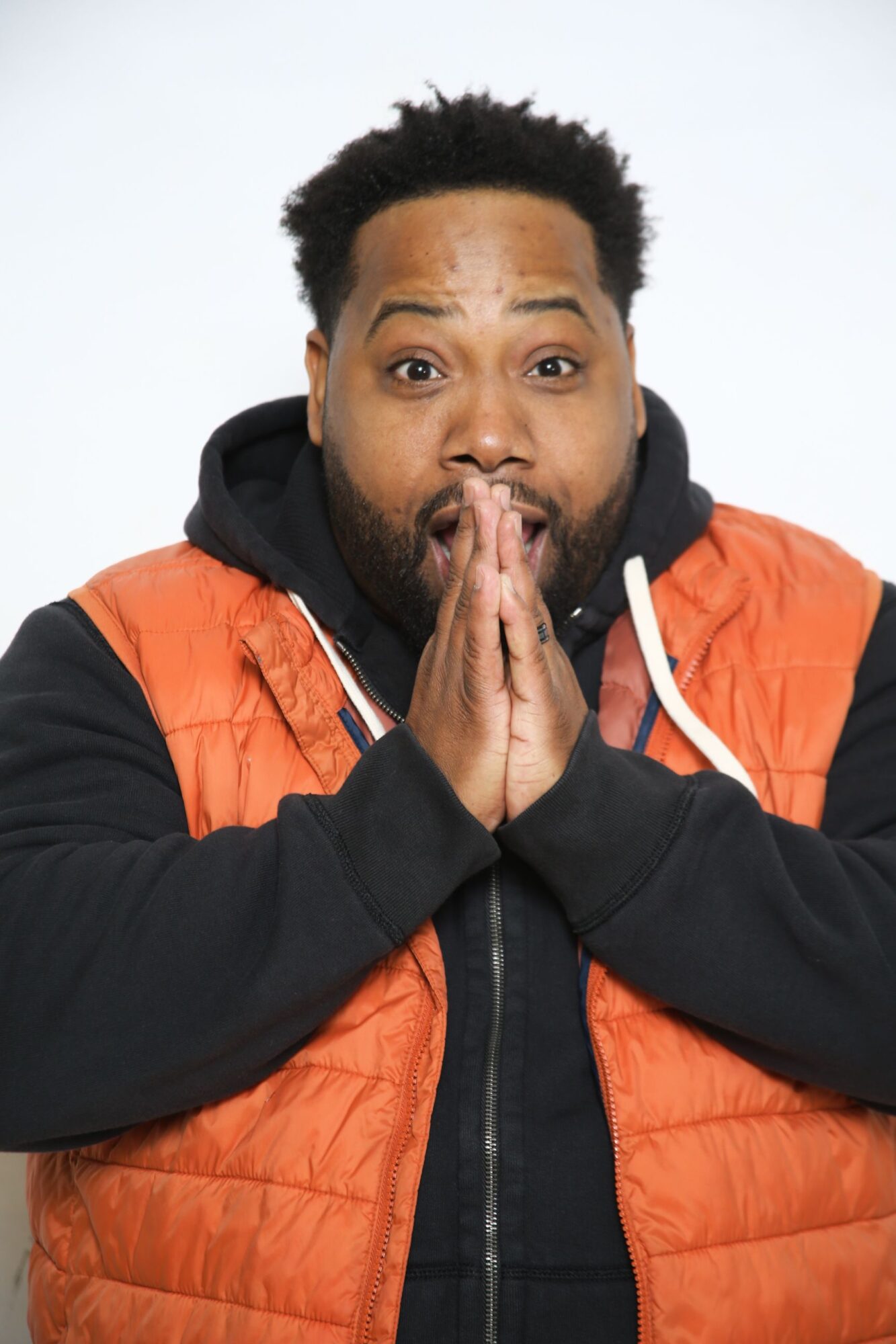 Person with hands pressed together near face, wearing an orange and black jacket, looking at camera.