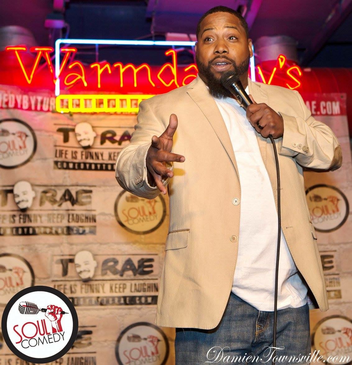 Man speaking into microphone on stage with colorful background and signs, wearing beige blazer and white shirt.