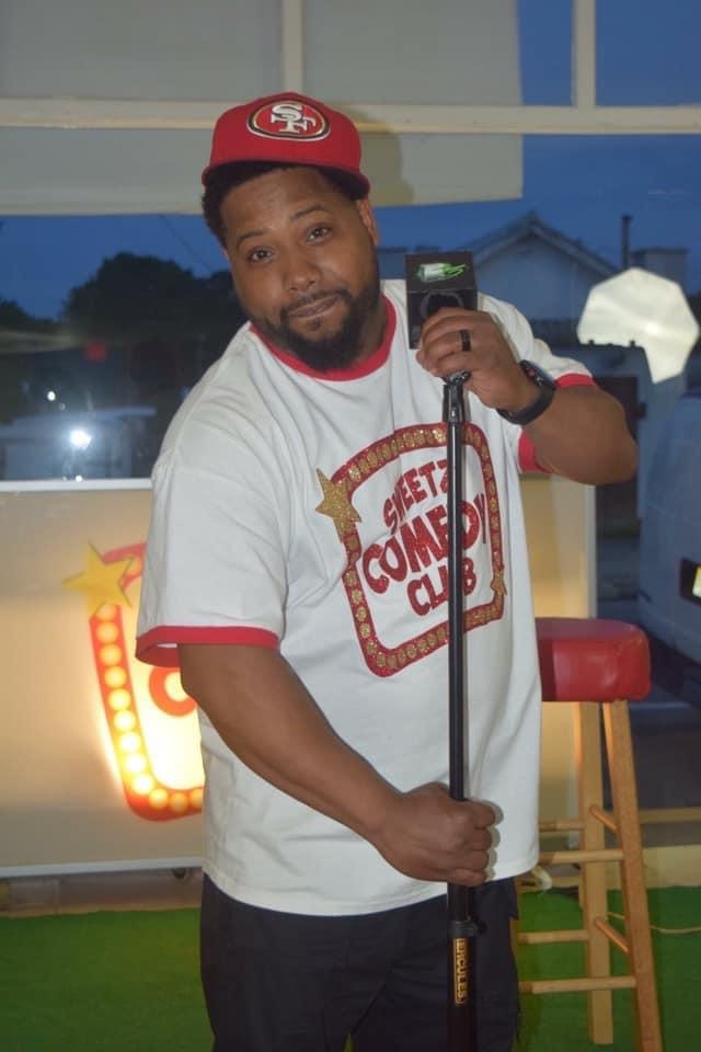 Man holding microphone, wearing a white T-shirt with red and gold design, red cap, standing indoors with window behind him.