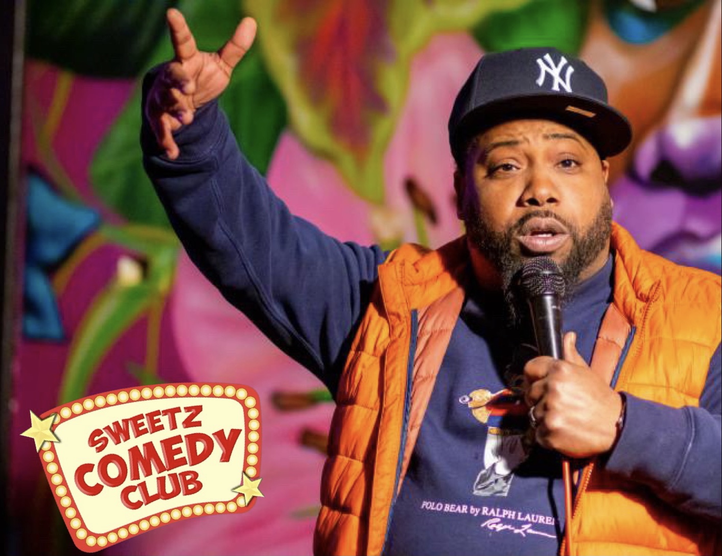 Comedian performing on stage with colorful background, holding microphone, wearing a cap and orange vest.