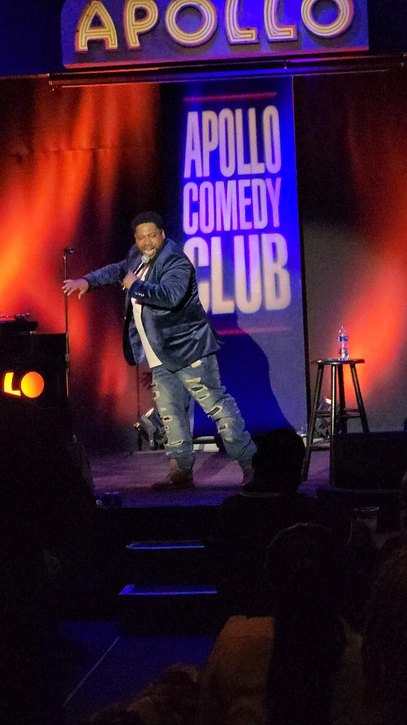 Comedian performing on stage at Apollo Comedy Club with a microphone, stage backdrop, and stool, in a dark theater setting.