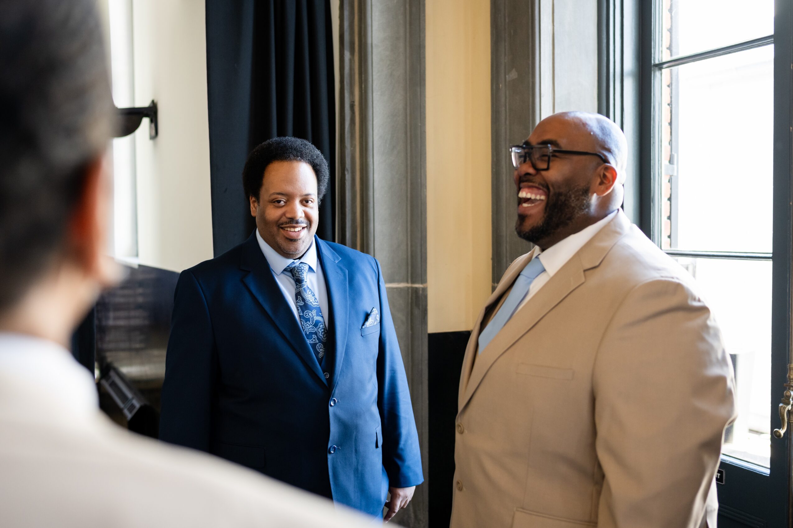 Two men in suits smiling and talking indoors near a window, with a third person partially visible in the foreground.
