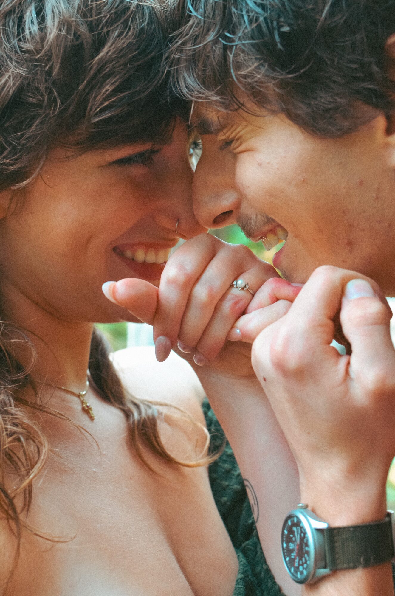 Two people with their faces close, smiling and touching hands, outdoors with blurred background