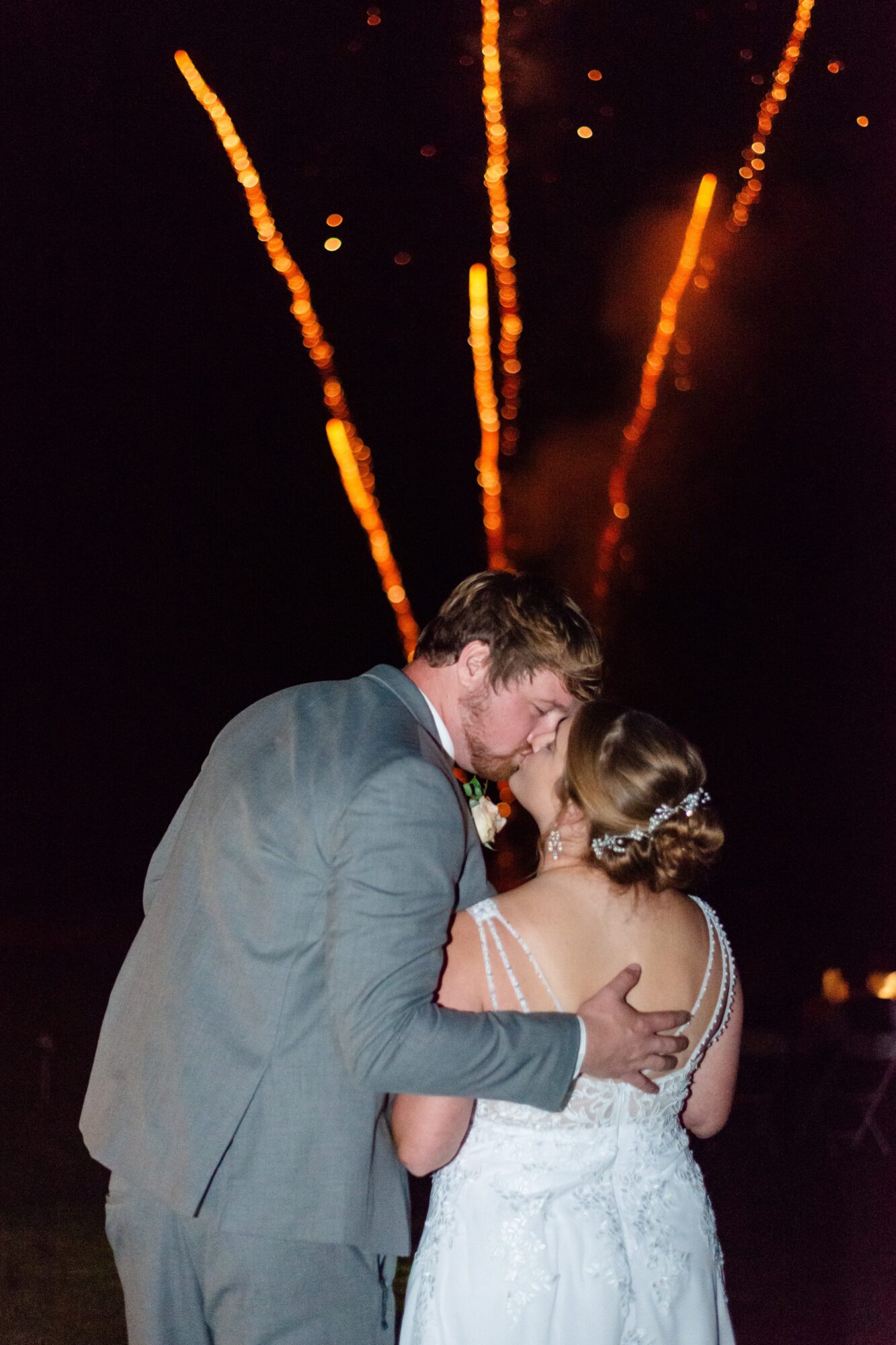 A couple in formal attire sharing a kiss at night with fireworks in the background.