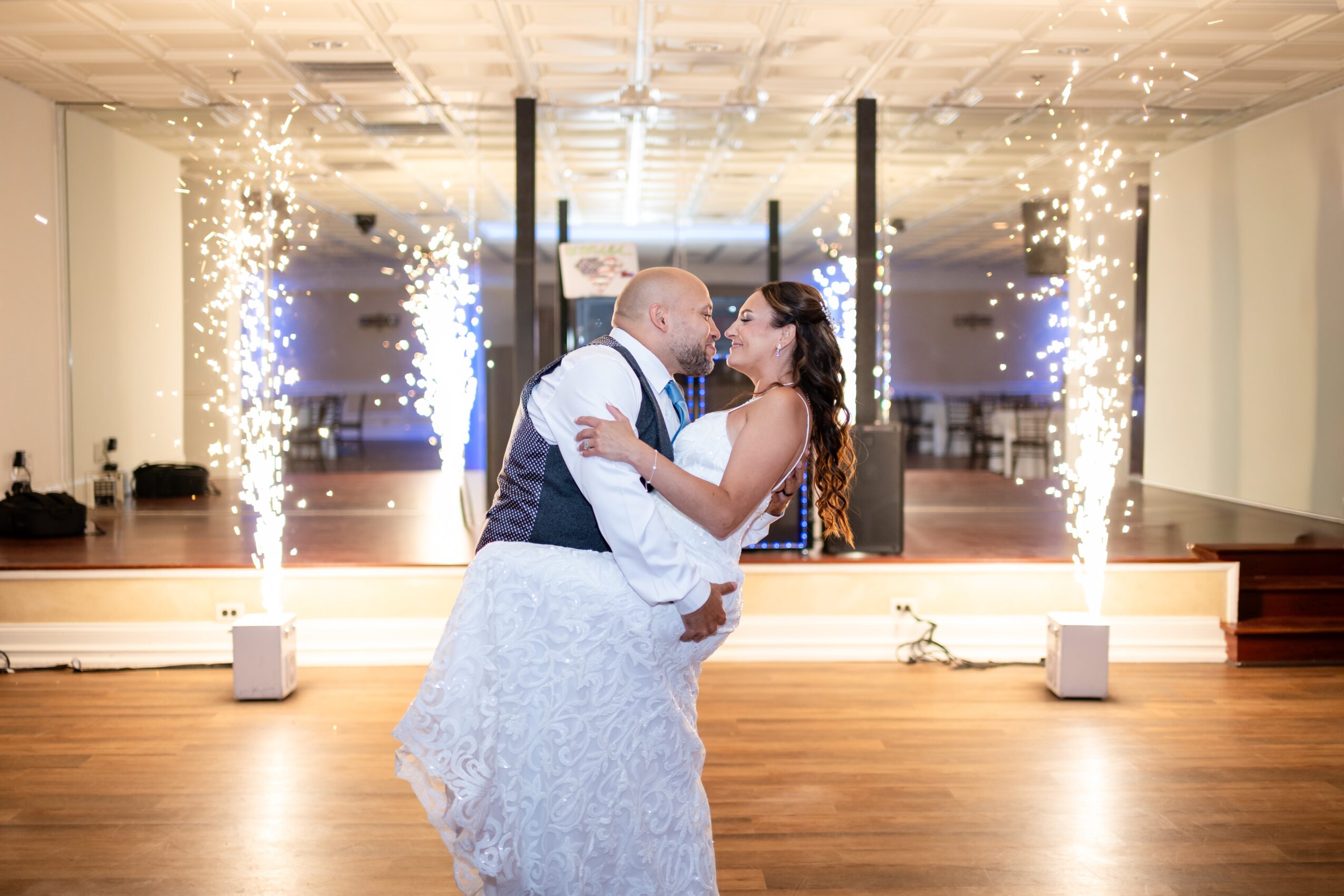 Couple dancing closely with fireworks in the background, celebrating indoors at a wedding reception, smiling and embracing.