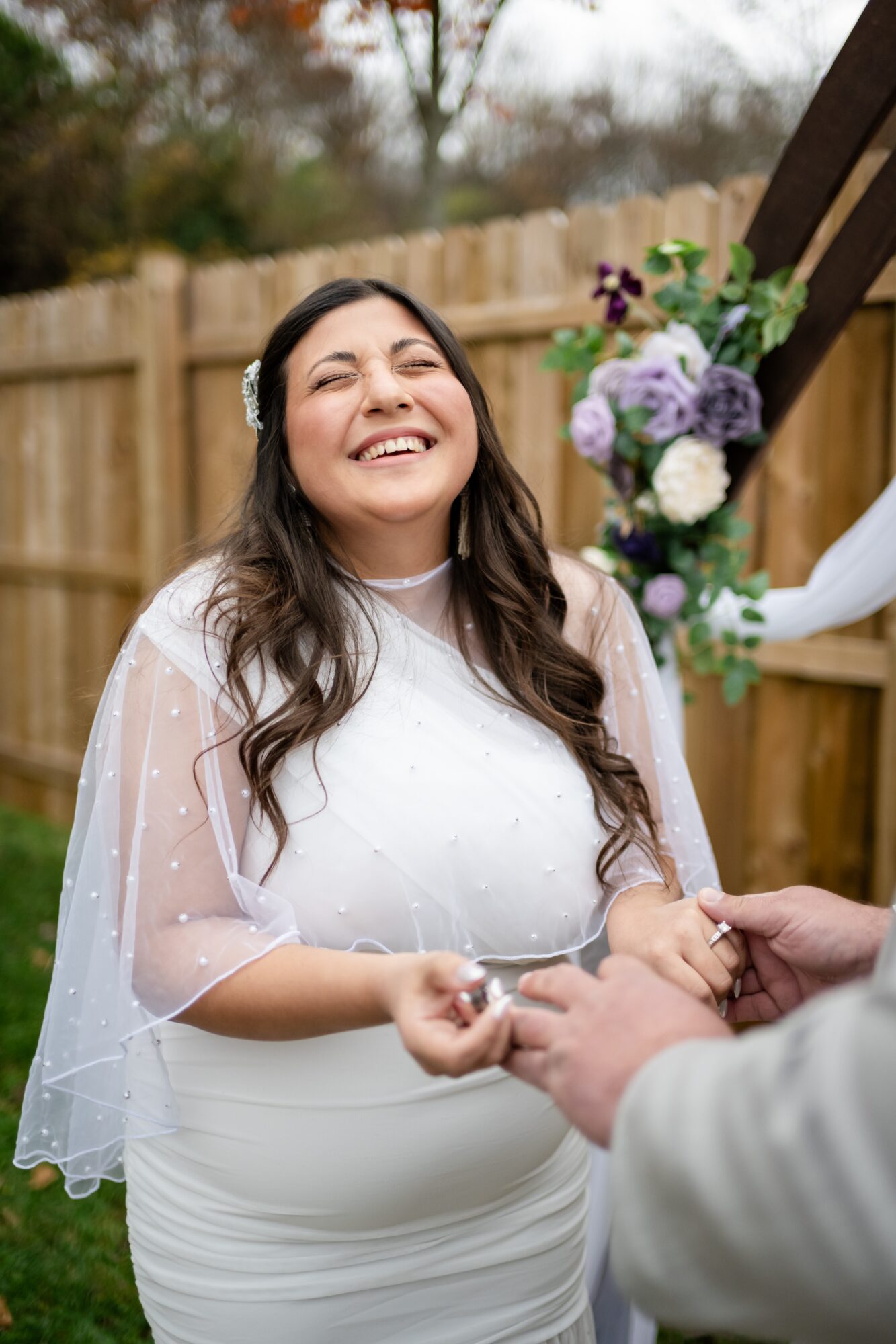 Woman in wedding dress smiling during outdoor ceremony, holding hands with partner, with floral arrangement nearby.