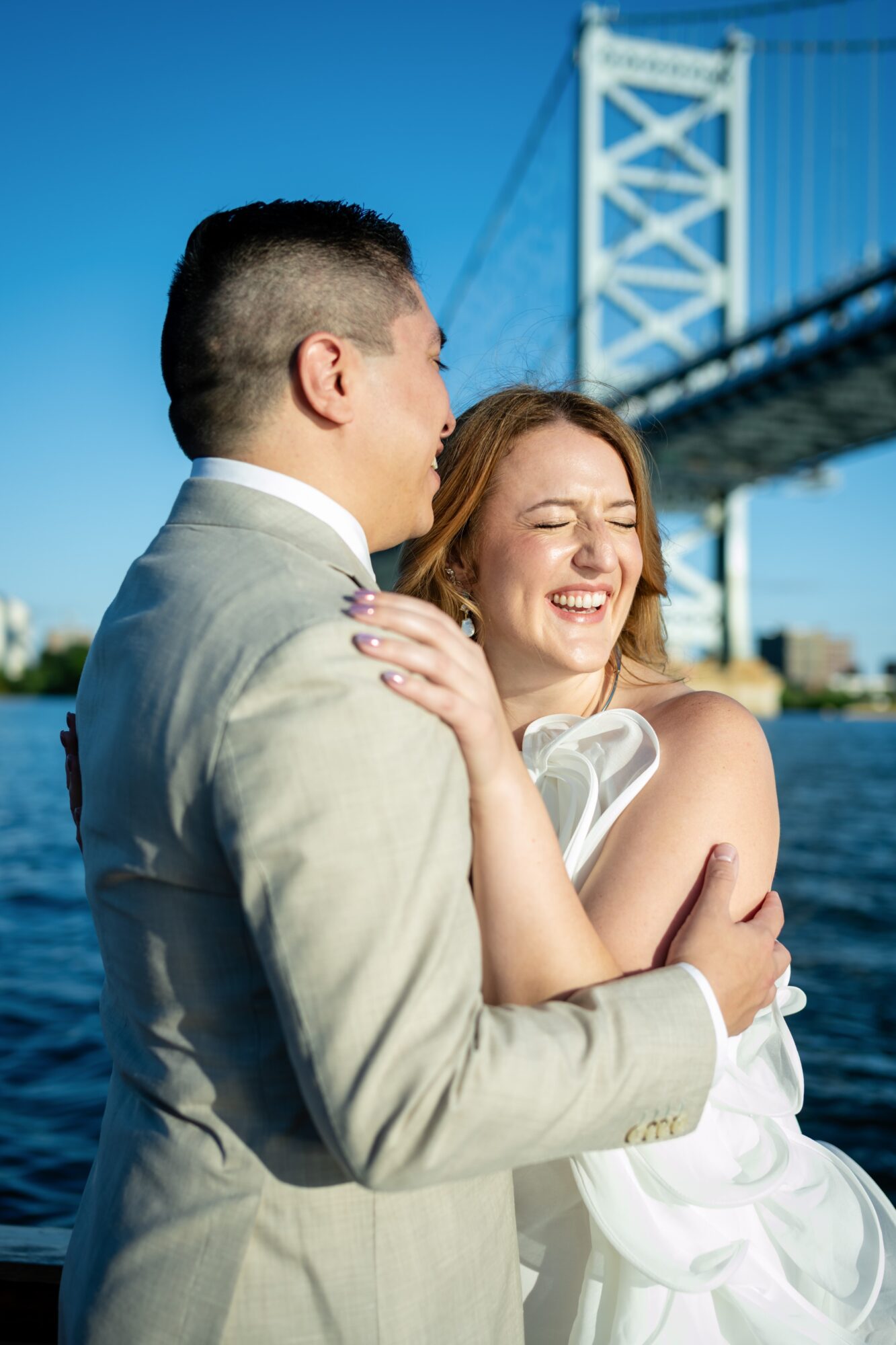 A smiling woman and a man in formal attire embrace outdoors with a bridge in the background.