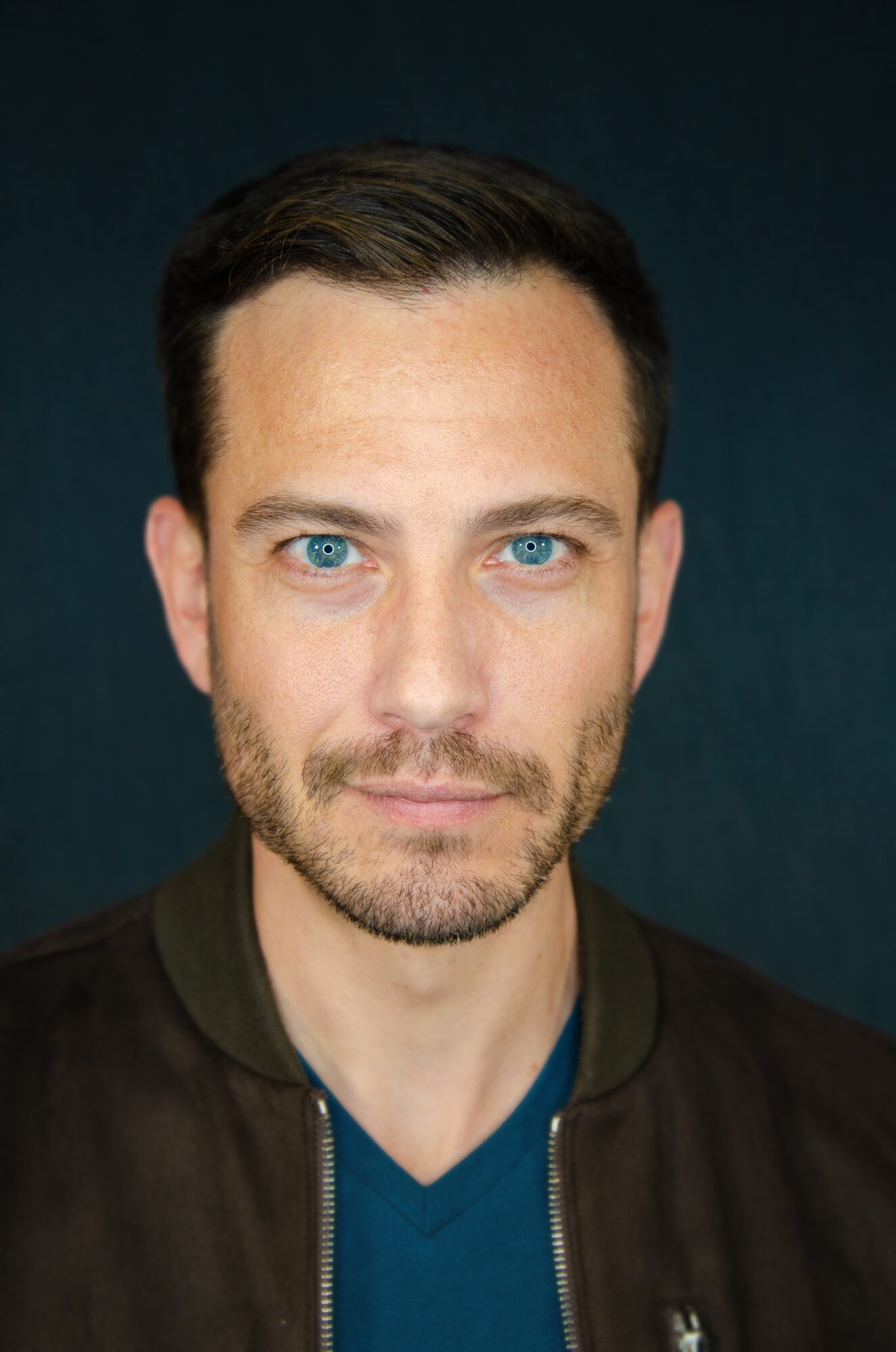 Portrait of a man with short dark hair, blue eyes, and a beard, wearing a dark jacket and blue shirt.
