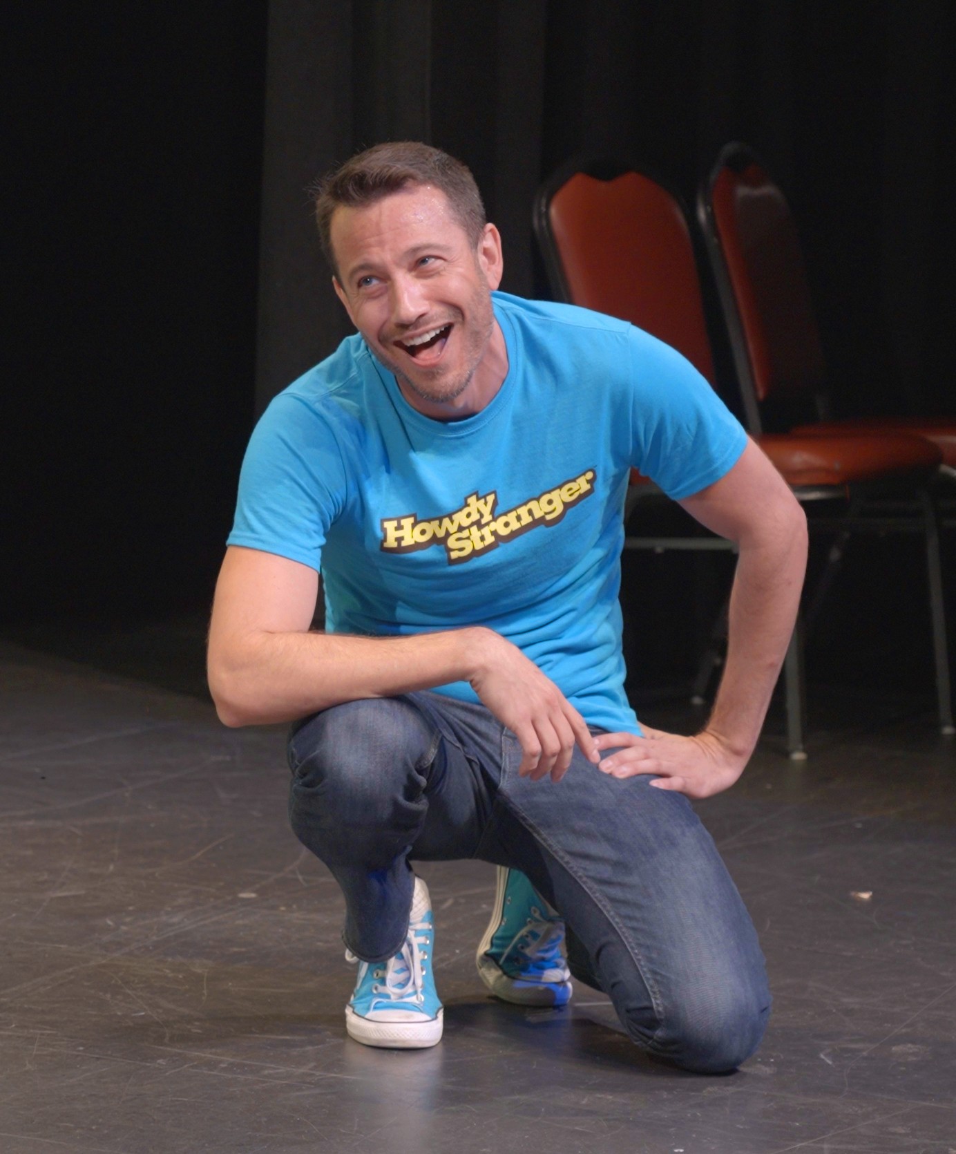 Man crouching on stage, smiling, wearing a blue t-shirt with yellow text, jeans, and sneakers, with chairs in the background.