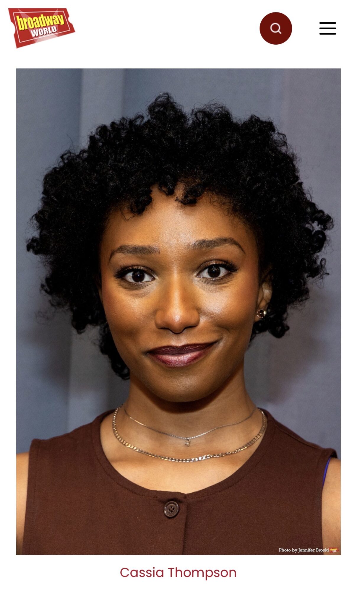 Portrait of a woman with short curly hair, wearing a brown top and a necklace, smiling at the camera.