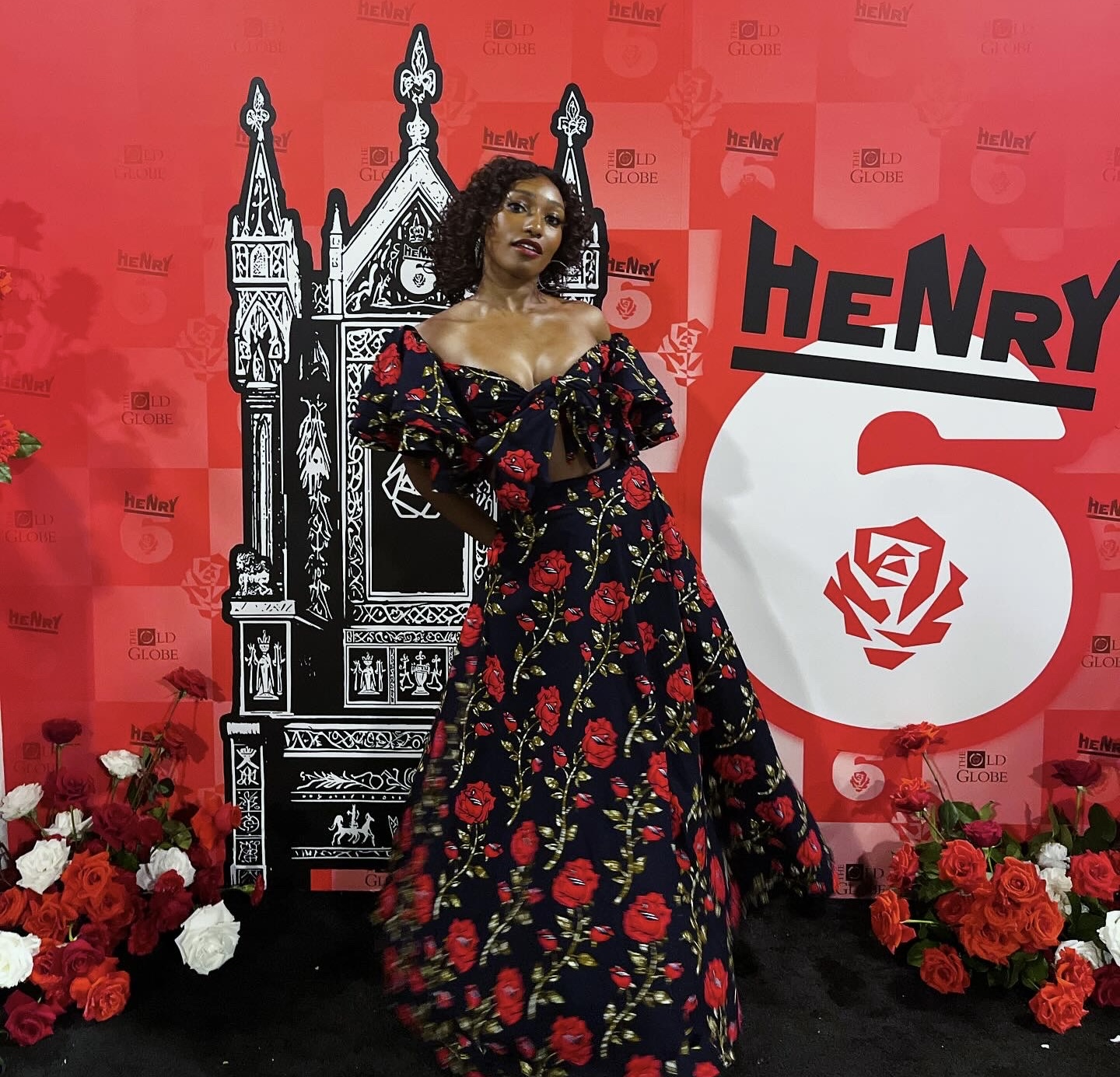 Woman in a floral gown standing in front of a red backdrop with floral arrangements and a black and white illustration.