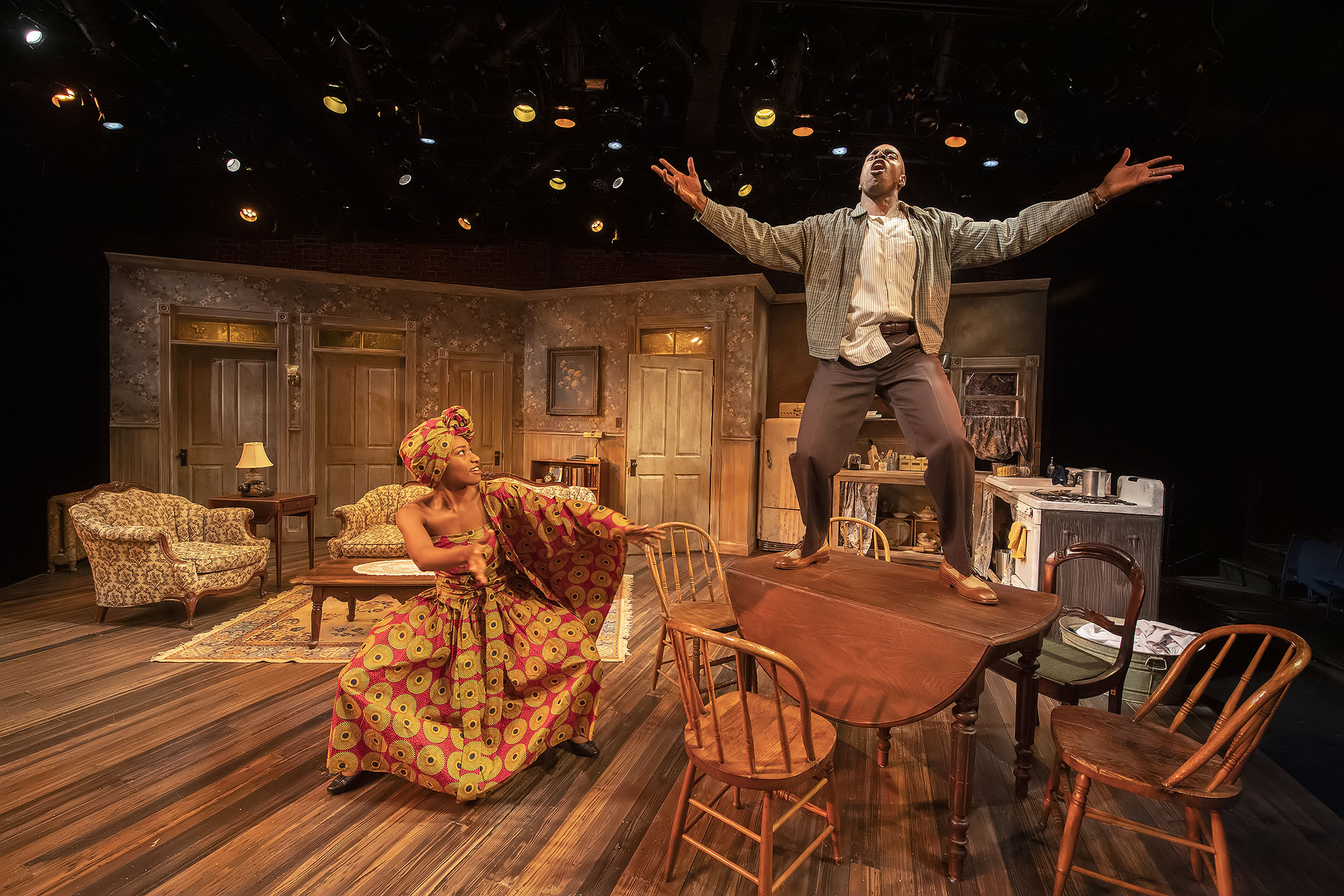 Stage scene with two actors, one standing with arms outstretched, the other sitting at a table, set in a cozy room.