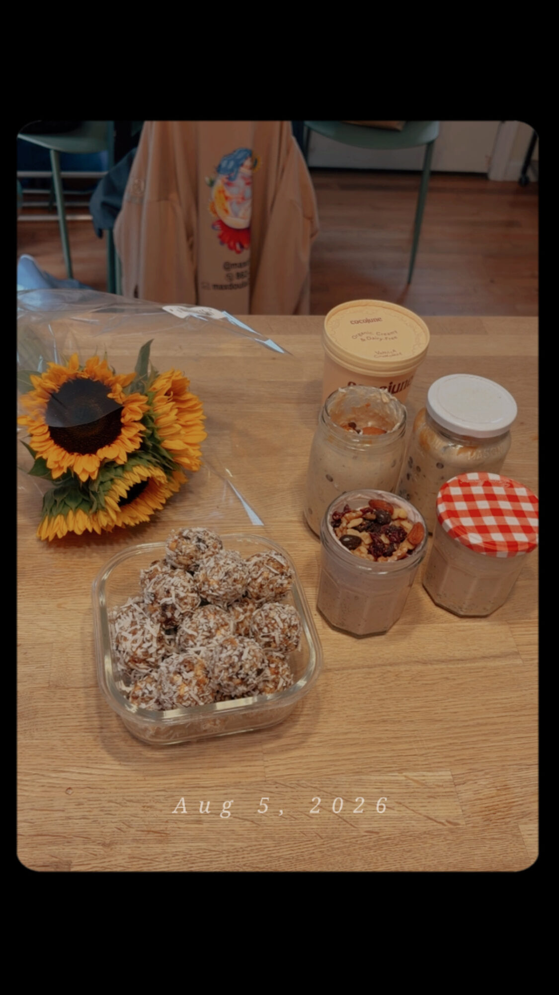 Table with sunflower bouquet, jars of baked goods, and a plastic container of cookies, with a chair and a person in the background.