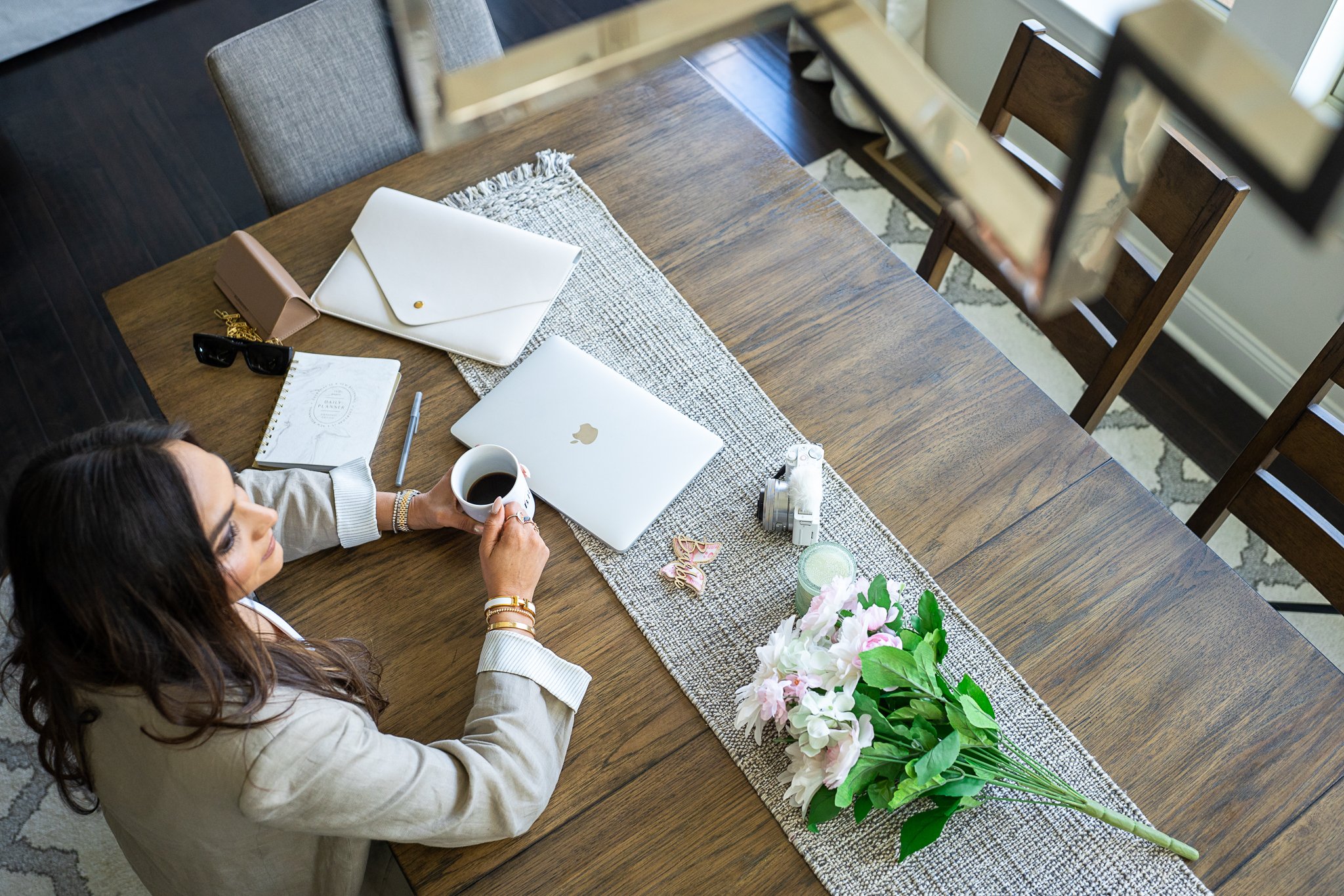 Woman sitting at a wooden table with a laptop, coffee, flowers, and papers, viewed from above.